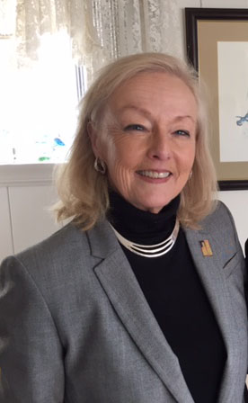 Sue Savary standing in front of window, smiling in gray jacket